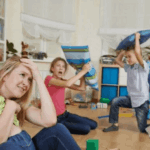 Stressed mother sitting on the floor holding a baby while children play loudly behind her in a busy living room