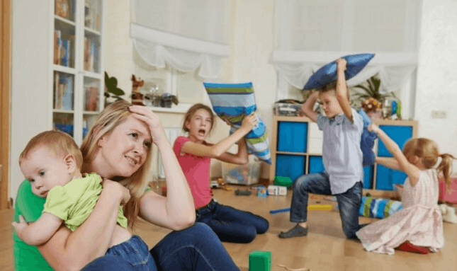 Stressed mother sitting on the floor holding a baby while children play loudly behind her in a busy living room