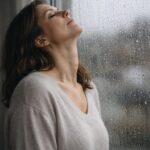 Woman standing beside a rain-covered window with her eyes closed and head tilted back, exhaling as she releases emotional weight during a season of uncertainty.